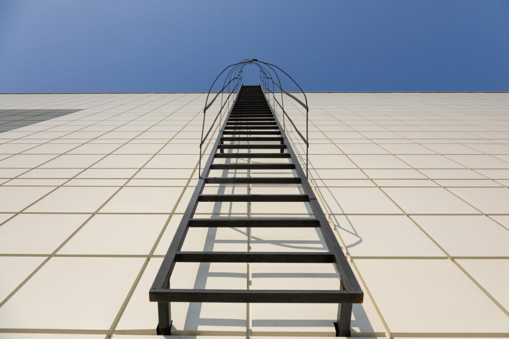 a long metal staircase on the gray modern facade of an industrial building, warehouse or shopping center against the blue sky. Symbol of the stairway to heaven